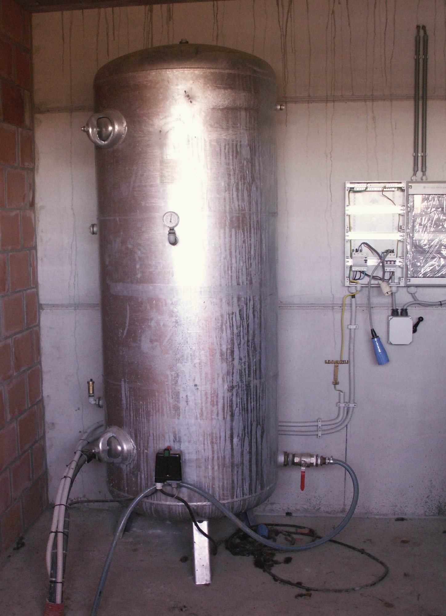 A tall stainless pressure tank in a pump room with gauges and wiring