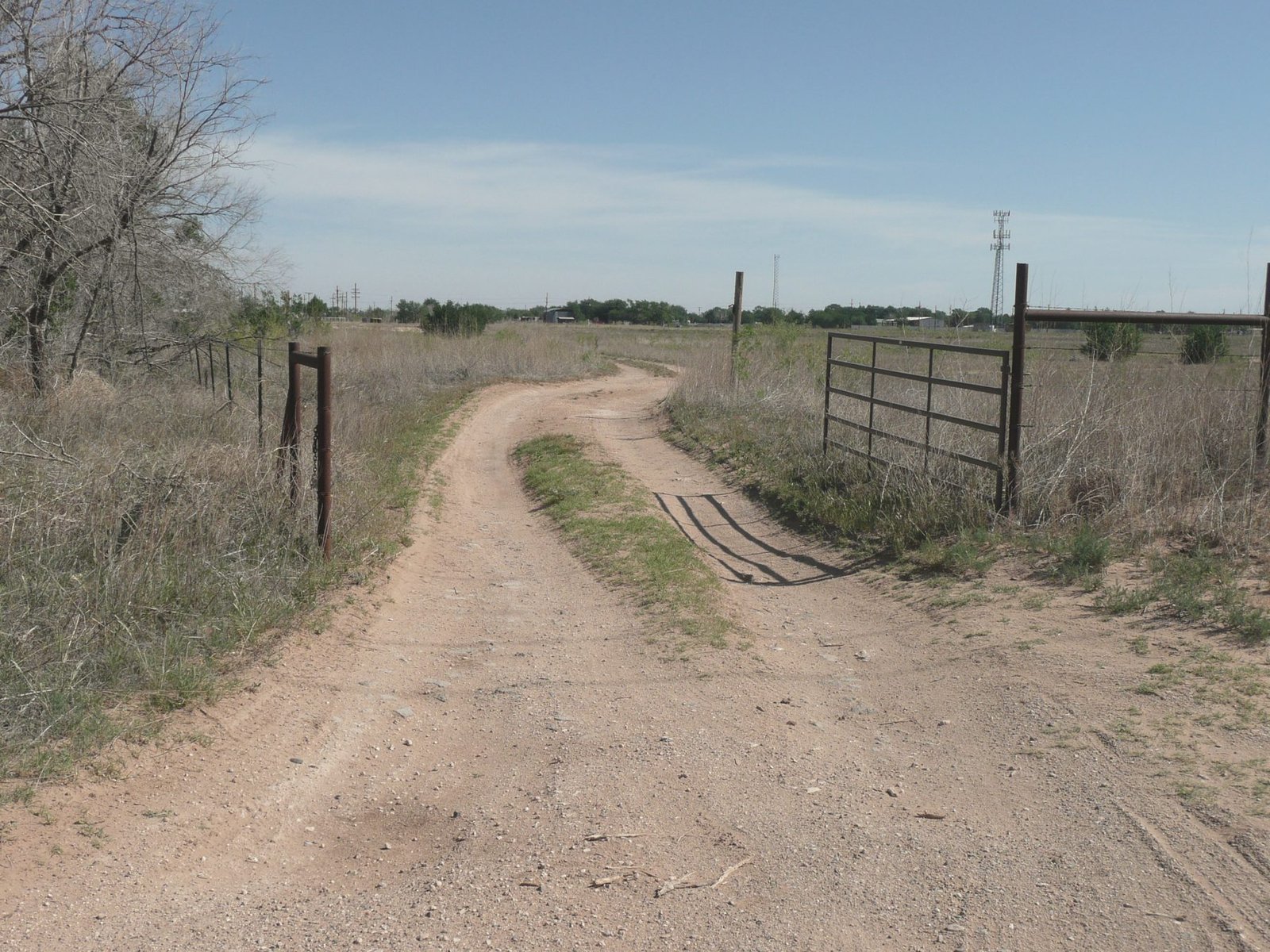 A dirt ranch road curving past an open pipe gate through dry brush