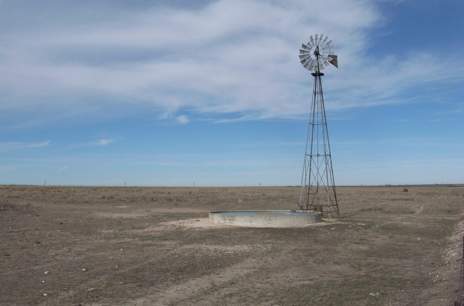 A windmill and a round stock tank on flat, dry plains under a pale sky
